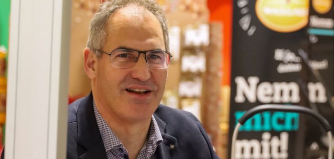 Landrat Diemtar Seefeldt sitzt in Hemd und Sakko an einer Supermarktkasse und scannt gerade eine Banane ab.