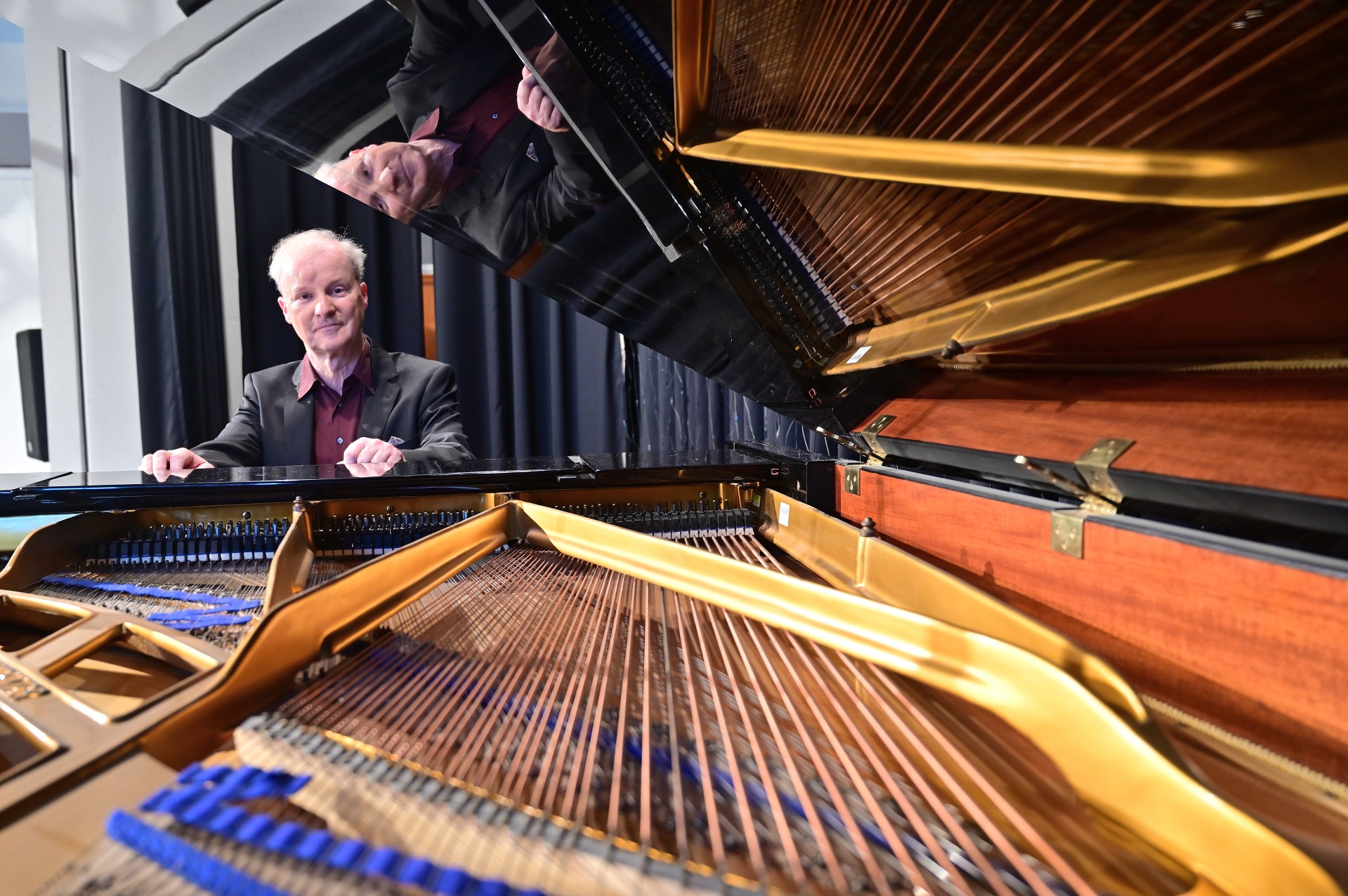 Der Pianist und Geiger Kolja Lessing sitzt an einem geöffneten Flügel. Der Blickwinkel zeigt das Innenleben des Instruments, mit den Saiten und der Mechanik im Vordergrund. Im Deckel des Flügels spiegelt sich sein Gesicht wider.