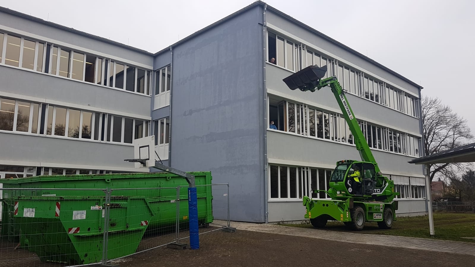 Außenaufnahme eines Schulgebäudes am PAMINA-Schulzentrum in Herxheim. Links vor dem Gebäude ist ein großer grüner Container für anfallenden Baustellenabfall zu sehen, rechts ein grünes Baustellenfahrzeug.