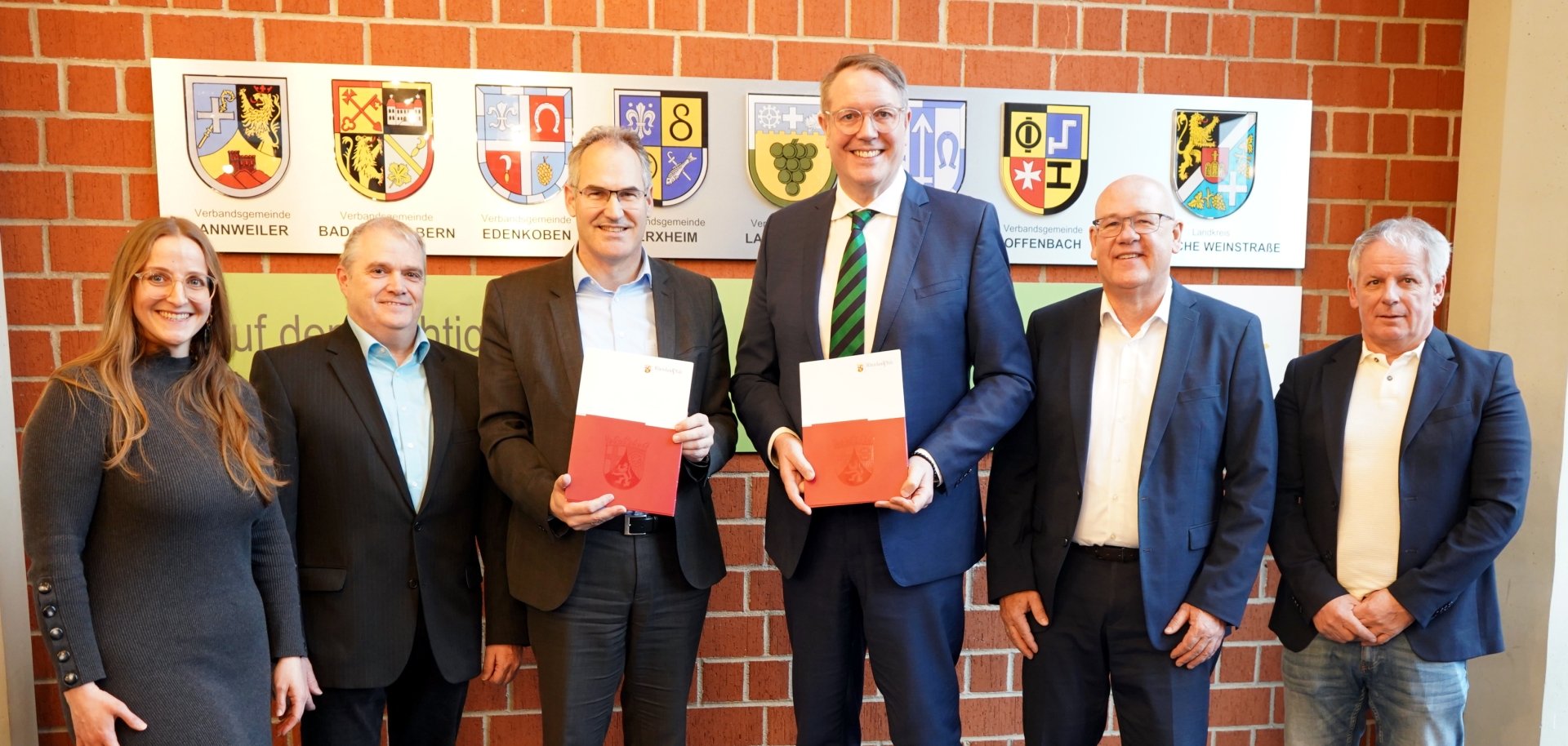 Gruppenbild im Kreissaal des Kreishauses SÜW. Von links nach rechts stehen Karolin Söltner, Sachbearbeiterin Breitbandausbau, Michael Stephany, Leiter des Referats Digitalisierung in der Kreisverwaltung SÜW, Landrat Dietmar Seefeldt, Ministerpräsident Alexander Schweitzer, MBB- Geschäftsführer Uwe König und Kreisbeigeordneter Werner Schreiner vor einer Backsteinwand zusammen, an der die Wappen des Landkreises SÜW sowie der sieben zugehörigen Verbandsgemeinden zu sehen sind. Landrat Seefeldt und Ministerpräsident Schweitzer präsentieren den übergebenen Bescheid.