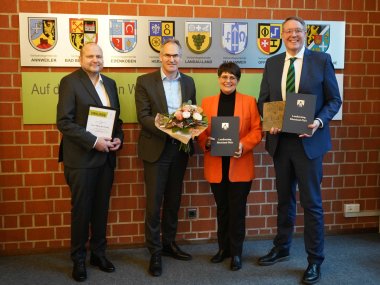 Von links nach rechts: Simon Bludovsky, Landrat Dietmar Seefeldt, Christine Schneider und Alexander Schweitzer. Foto: KV SÜW Simon Bludovsky, Landrat Dietmar Seefeldt, Christine Schneider und Alexander Schweitzer stehen im Kreishaus der Kreisverwaltung Südliche Weinstraße vor einer Backsteinwand. Darauf sind im Hintergrund das Wappen des Landkreises sowie der zugehörigen Verbandsgemeinde zu sehen.