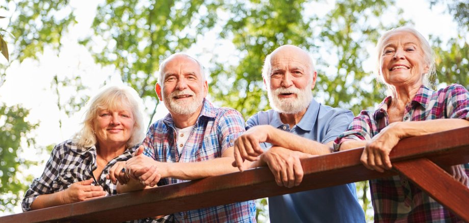 Vier Seniorinne und Senioren lehnen sich auf ein hölzernes Geländer und lächeln den Betrachter an. Im Hintergrund sind unscharf grüne Laubbäume zu sehen.