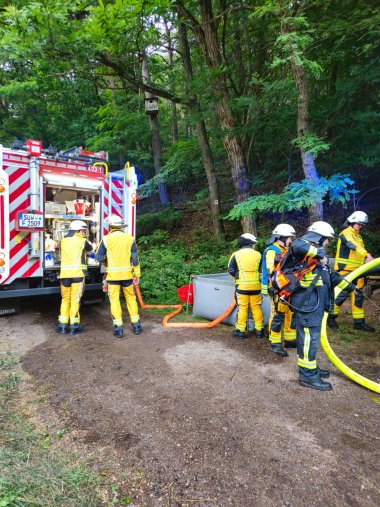 Mehrere Feuerwehrleute in gelber Schutzausrüstung stehen neben einem Feuerwehrauto im Wald. Sie bereiten Schläuche vor. Im Hintergrund dichter Wald mit grünen Bäumen.