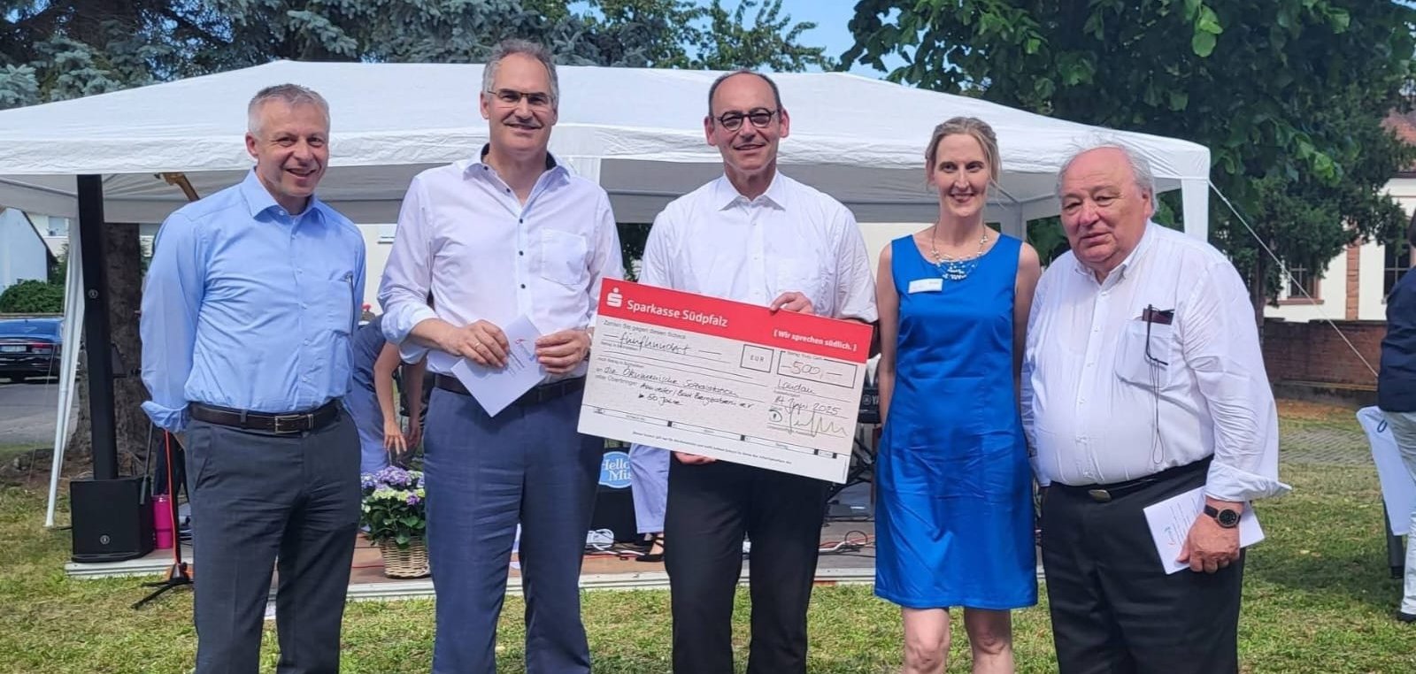 Landrat Dietmar Seefeldt und die Verantwortlichen der Ökumenischen Sozialstation stehen in einer Reihe vor einem weißen Pavillon. Der Mann in der Mitte hält einen Spendenscheck in der Hand.