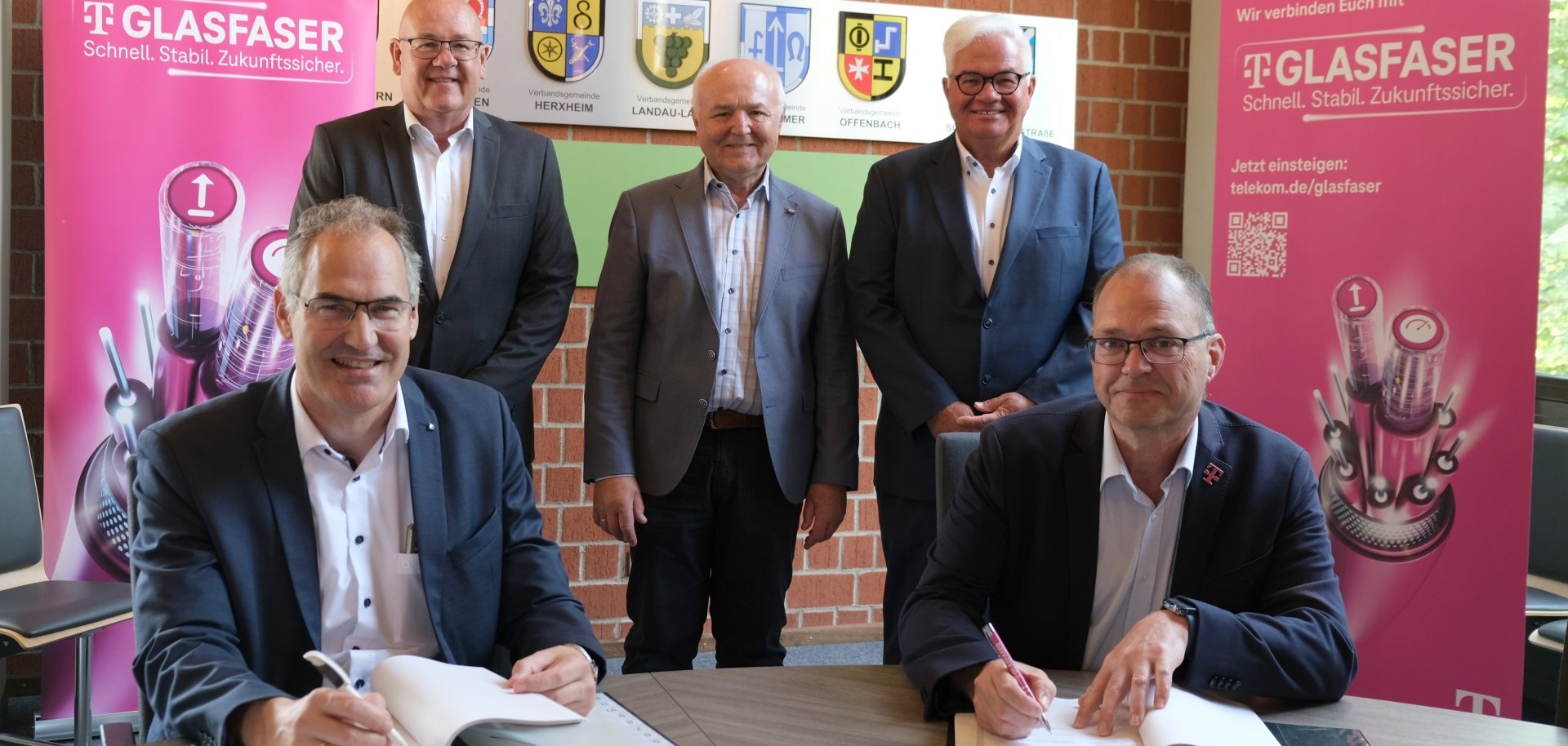 Gruppenbild: Landrat Dietmar Seefeldt, Uwe König, Albert Schädler, Gerd Schäfer und Martin Stiebitz bei der Vertragsunterzeichnung in der Kreisverwaltung. Seefeldt und Stiebitz sind – an einem Tisch sitzend, mit Kugelschreiber in der Hand und vor ihnen liegenden Vertragsunterlagen – am linken und rechten Bildrand zu sehen. Die anderen Männer stehen dazwischen. Im Hintergrund sind magentafarbene Rollups der Telekom zu sehen.