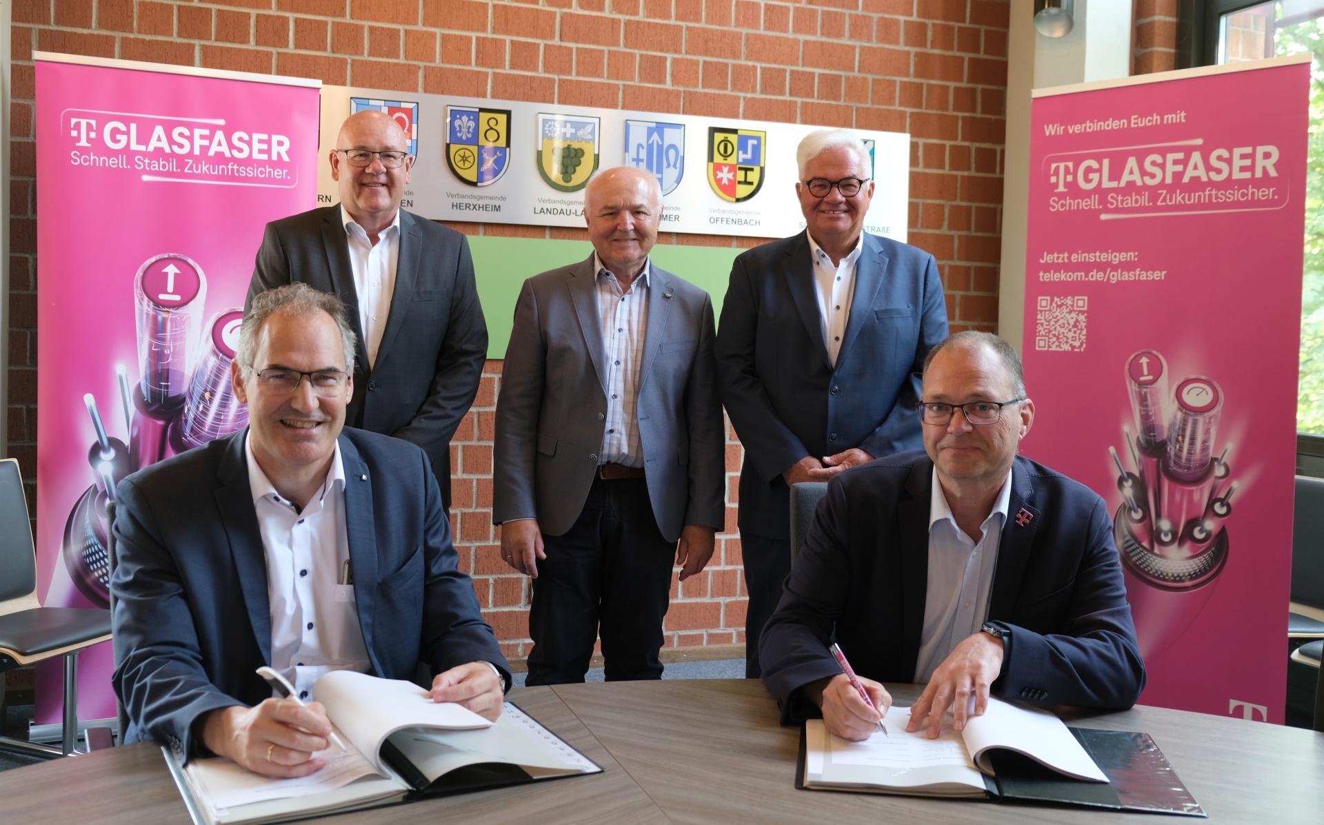 Gruppenbild: Landrat Dietmar Seefeldt, Uwe König, Albert Schädler, Gerd Schäfer und Martin Stiebitz bei der Vertragsunterzeichnung in der Kreisverwaltung. Seefeldt und Stiebitz sind – an einem Tisch sitzend, mit Kugelschreiber in der Hand und vor ihnen liegenden Vertragsunterlagen – am linken und rechten Bildrand zu sehen. Die anderen Männer stehen dazwischen. Im Hintergrund sind magentafarbene Rollups der Telekom zu sehen.