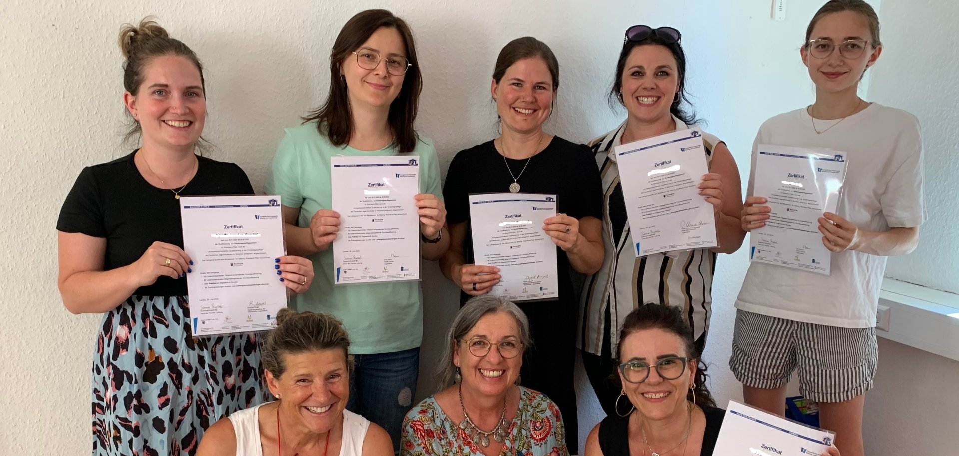 Gruppenfoto: Acht Frauen stehen beziehungsweise knien in zwei Reihen vor einer weißen Wand und halten ihr Zertifikat zum absolvierten Kurs zur Tagespflegeperson in Händen.