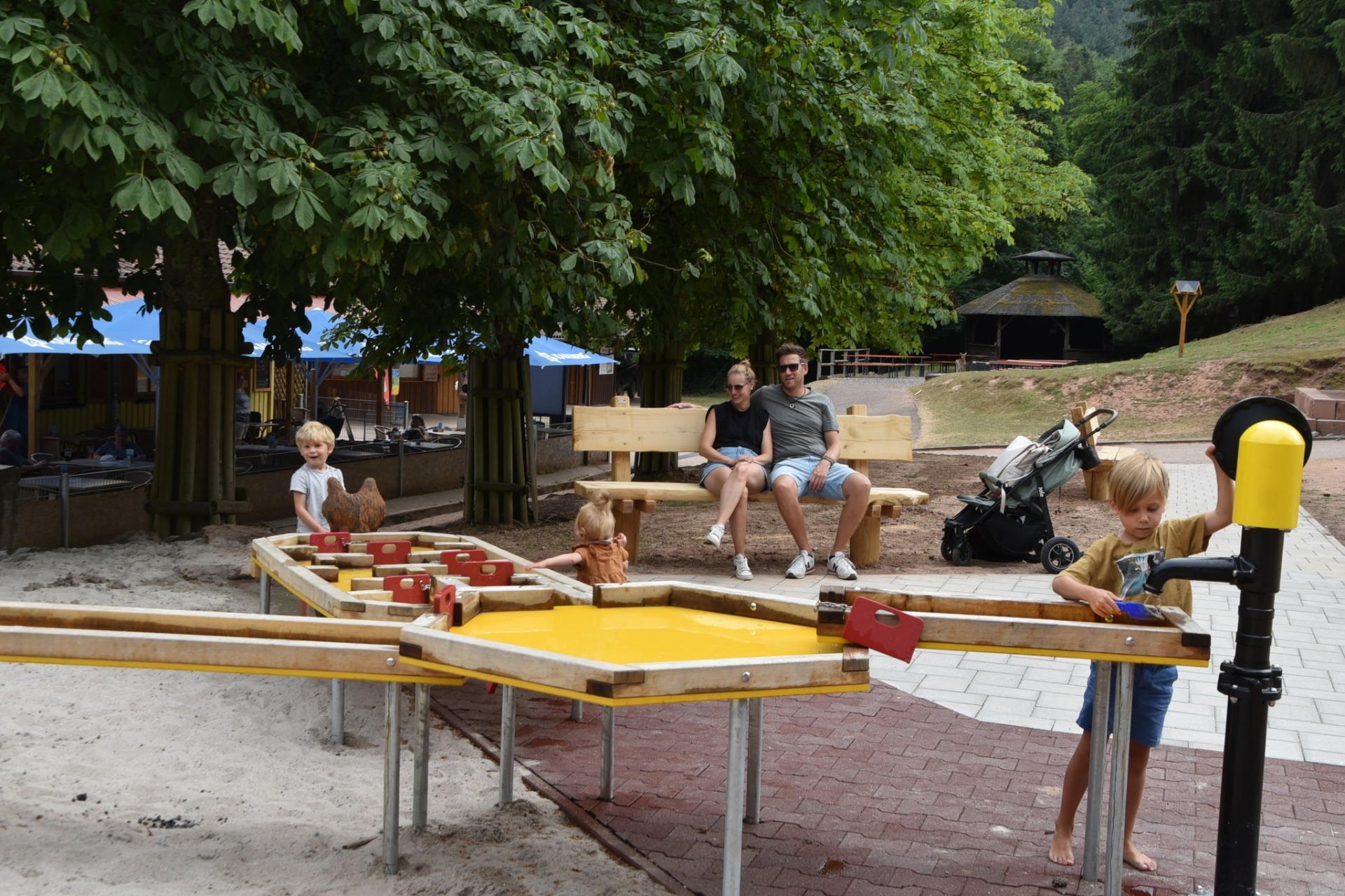 Drei Kinder spielen an einem großen Matschtisch im Wild- und Wanderpark Südliche Weinstraße. Die Eltern, ein junger Mann und eine junge Frau in sommerlicher Kleidung, sitzen im Hintergrund auf einer Holzbank und sehen den Kindern zu. Das Areal ist umgeben von Bäumen, im Hintergrund sieht man außerdem blaue Sonnenschirme und einen Sitzbereich der Gastronomie im Park.