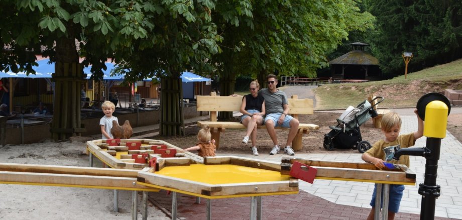 Drei Kinder spielen an einem großen Matschtisch im Wild- und Wanderpark Südliche Weinstraße. Die Eltern, ein junger Mann und eine junge Frau in sommerlicher Kleidung, sitzen im Hintergrund auf einer Holzbank und sehen den Kindern zu. Das Areal ist umgeben von Bäumen, im Hintergrund sieht man außerdem blaue Sonnenschirme und einen Sitzbereich der Gastronomie im Park.