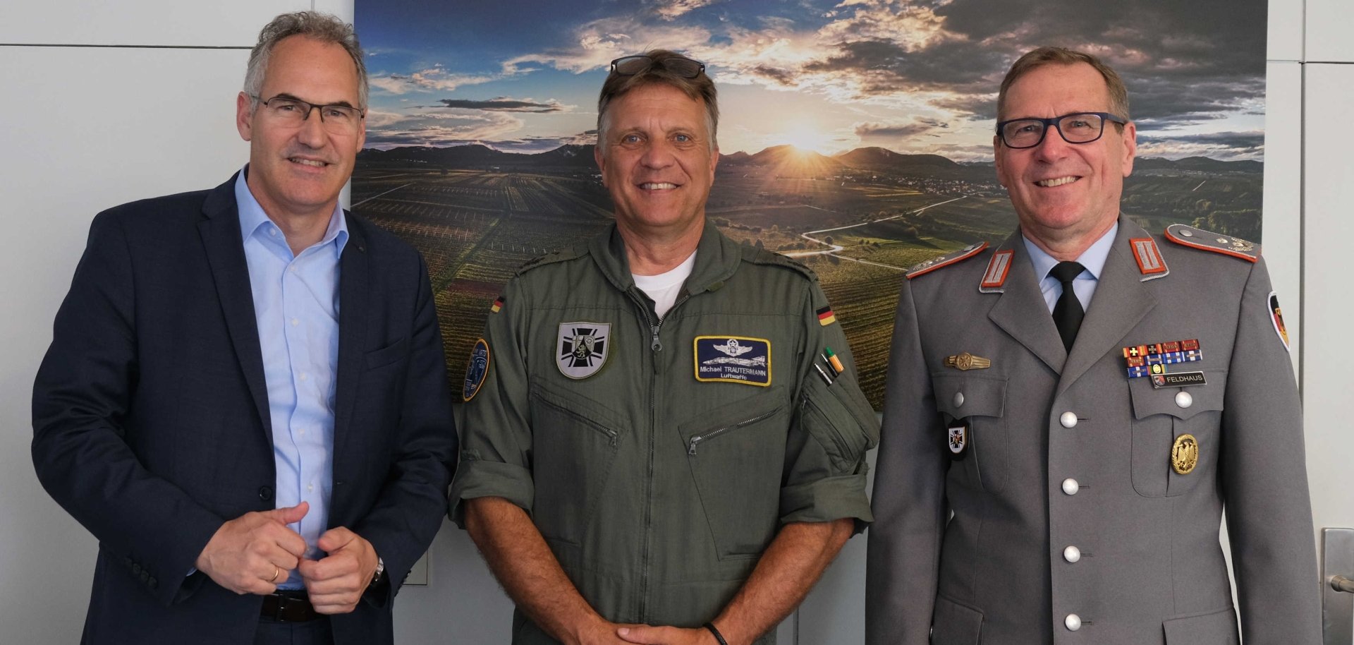 Drei Männer stehen nebeneinander und lächeln in die Kamera. Links ein Mann im Anzug, in der Mitte ein Mann in grüner Uniform mit Abzeichen der Bundeswehr, rechts ein Mann in grauer Bundeswehruniform. Im Hintergrund ist ein Landschaftsfoto mit Sonnenuntergang zu sehen.