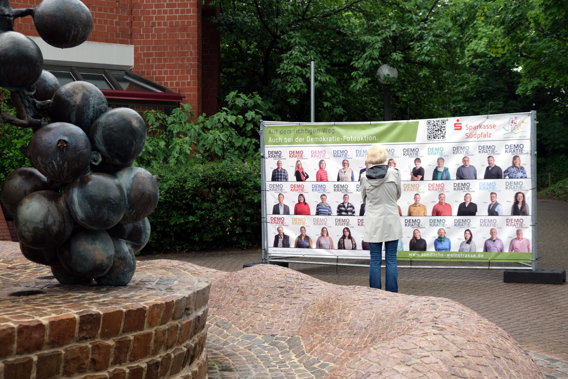 Eine Person betrachtet eines der Bauzaunbanner mit Fotos von Menschen, die sich im Rahmen der Fotoaktion für die Demokratie haben ablichten lassen. Das Banner steht vor dem Kreishaus SÜW in Landau. Am linken Bildrand ist im Vordergrund ein Teil des Früchtebrunnens vor dem Kreishaus zu sehen – bronzene Trauben.