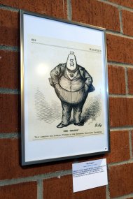 Mehr als 40 Werke von Thomas Nast, die meisten mit Bezug zum Thema Demokratie, sind aktuell im Kreishaus zu sehen. Foto: KV SÜW Die Karikatur "The Brains" von Thomas Nast hängt gerahmt an einer Backsteinwand im Kreishaus. Sie zeigt eine völlige Person in einem dreiteiligen Anzug, die anstelle eines Kopfes einen Sack mit Dollarzeichen hat.