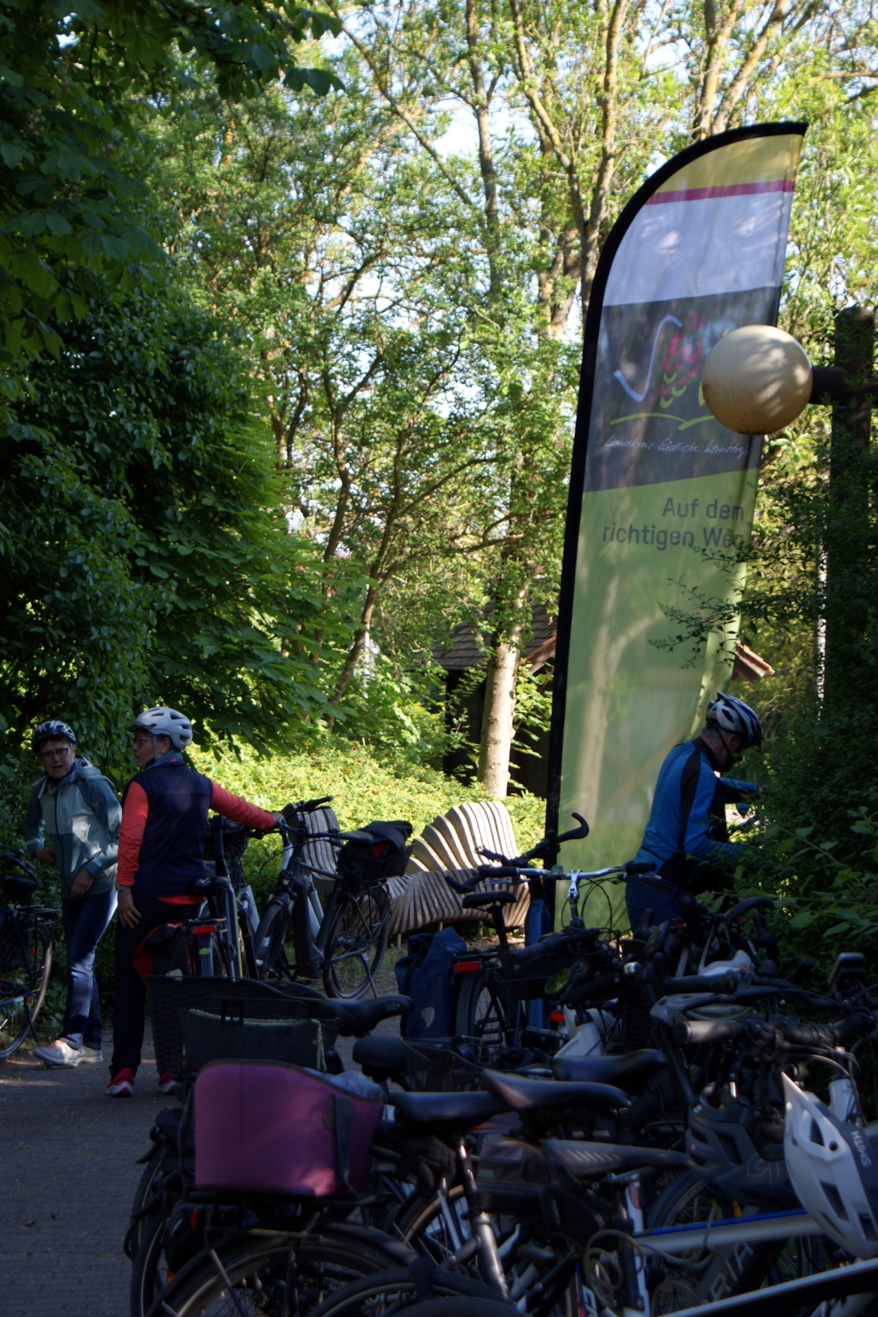 Mehrere abgestellte Fahrräder stehen vor einer Beachflag mit dem Logo der Kreisverwaltung Südliche Weinstraße. Im Hintergrund zwei Frauen und ein Mann an ihren Fahrrädern.