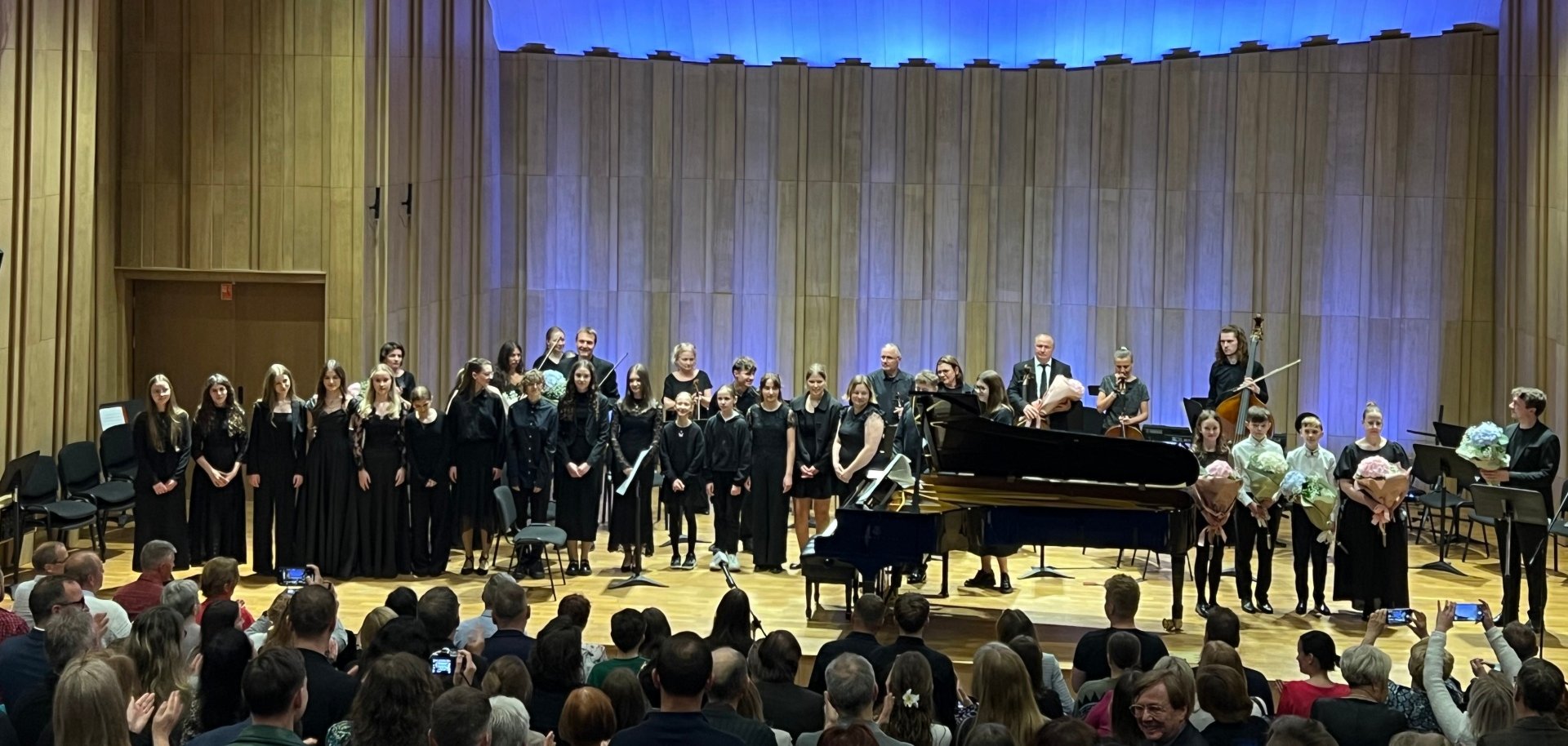 Das Ensemble aus polnischen Schülerinnen und Schüler und Lehrkräften der drei Musikschulen aus dem Landkreis SÜW, Oswiecim und Wissembourg auf der Bühne. Ein Publikum vor der Bühne gibt Beifall und fotografiert mit Mobiltelefonen. Einige Personen auf der Bühne haben Blumensträuße in der Hand.