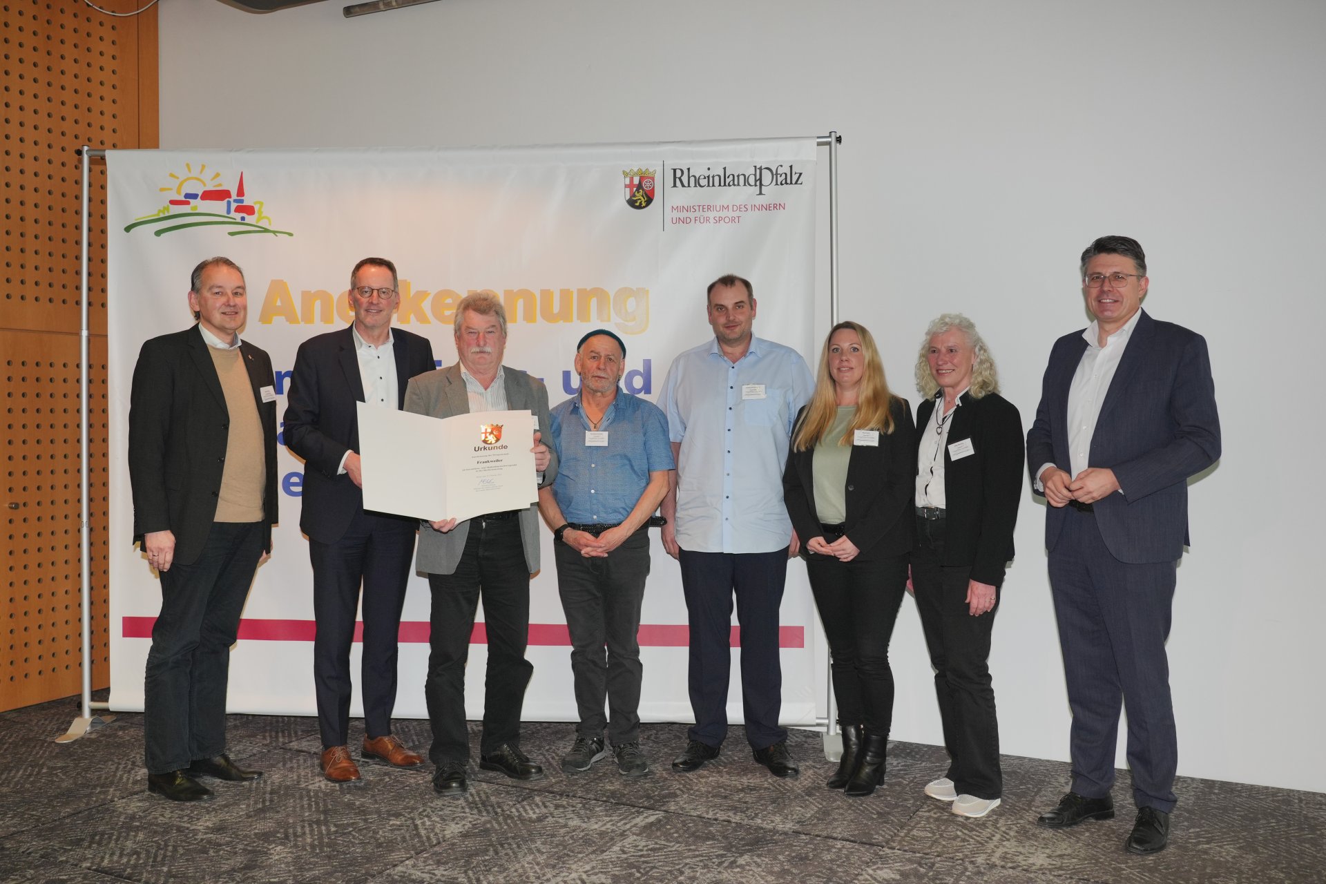Acht Personen stehen nebeneinander in einem Veranstaltungsraum und blicken in die Kamera. Eine Person in der Mitte hält eine große Urkunde mit dem Wappen von Rheinland-Pfalz. Im Hintergrund ist ein Banner mit dem Logo des rheinland-pfälzischen Innenministeriums sowie Schriftzügen zur Ehrung von Gemeinden zu sehen.