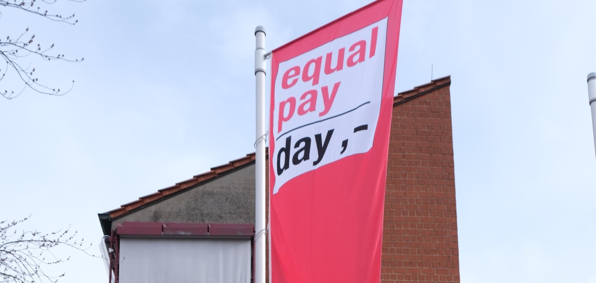 Rote Fahne mit der Aufschrift „Equal Pay Day“ an einem Fahnenmast vor einem Backsteingebäude. Die Flagge symbolisiert den Aktionstag für gleiche Bezahlung von Frauen und Männern.