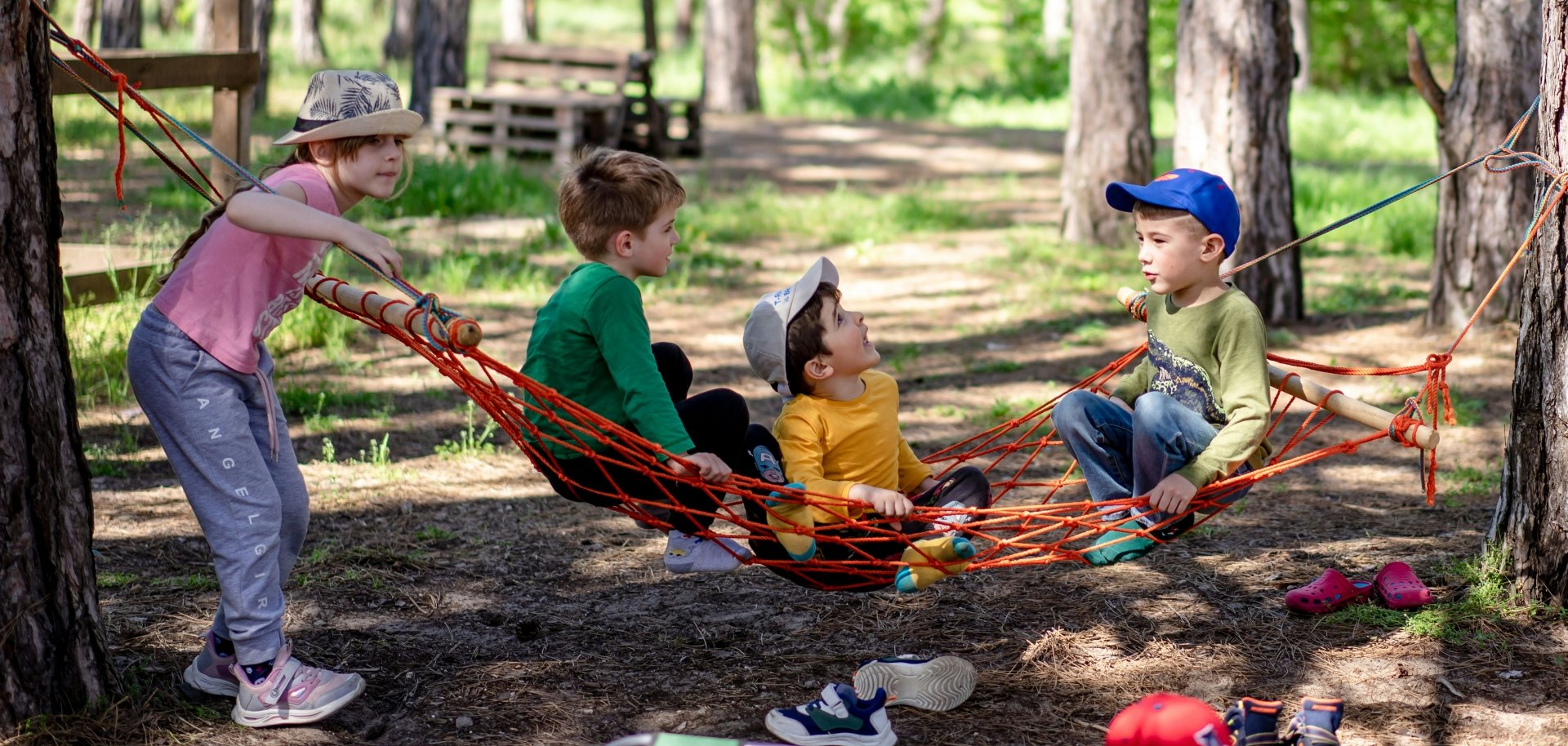 Vier Kinder spielen in einem Waldgebiet mit einer roten Hängematte, die zwischen zwei Bäumen gespannt ist. Drei Kinder sitzen in der Hängematte, während ein Mädchen sie festhält. Sie tragen bunte Kleidung und verschiedene Kopfbedeckungen. Im Hintergrund sind weitere Bäume und eine Holzkonstruktion zu sehen. Die Szene wirkt fröhlich und sommerlich.