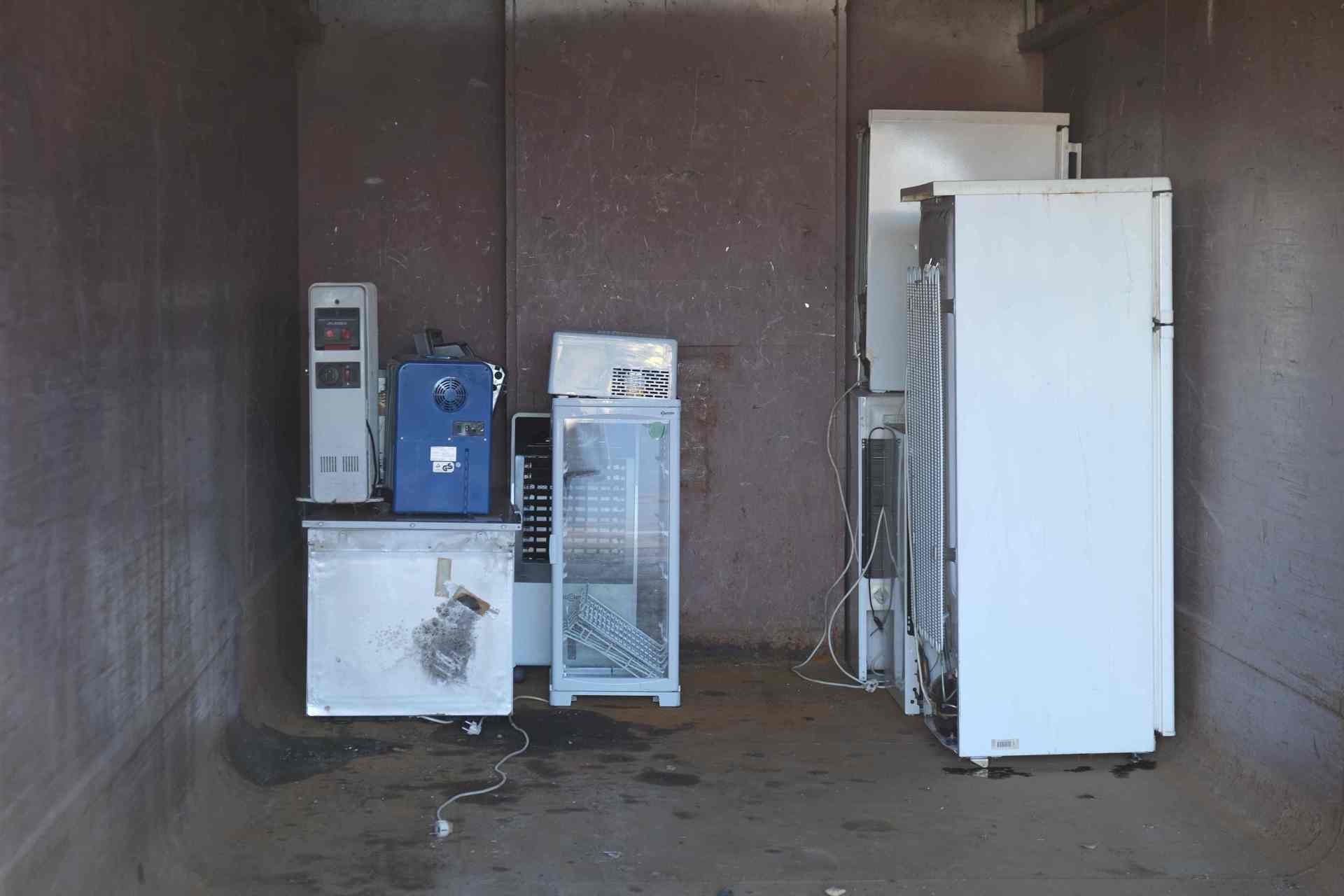 Mehrere ausgediente Elektrogeräte: Zu sehen sind unter anderem alte Kühlschränke, ein Gefrierschrank, ein Getränkekühler sowie weitere elektronische Geräte. Einige Kabel hängen lose herunter.