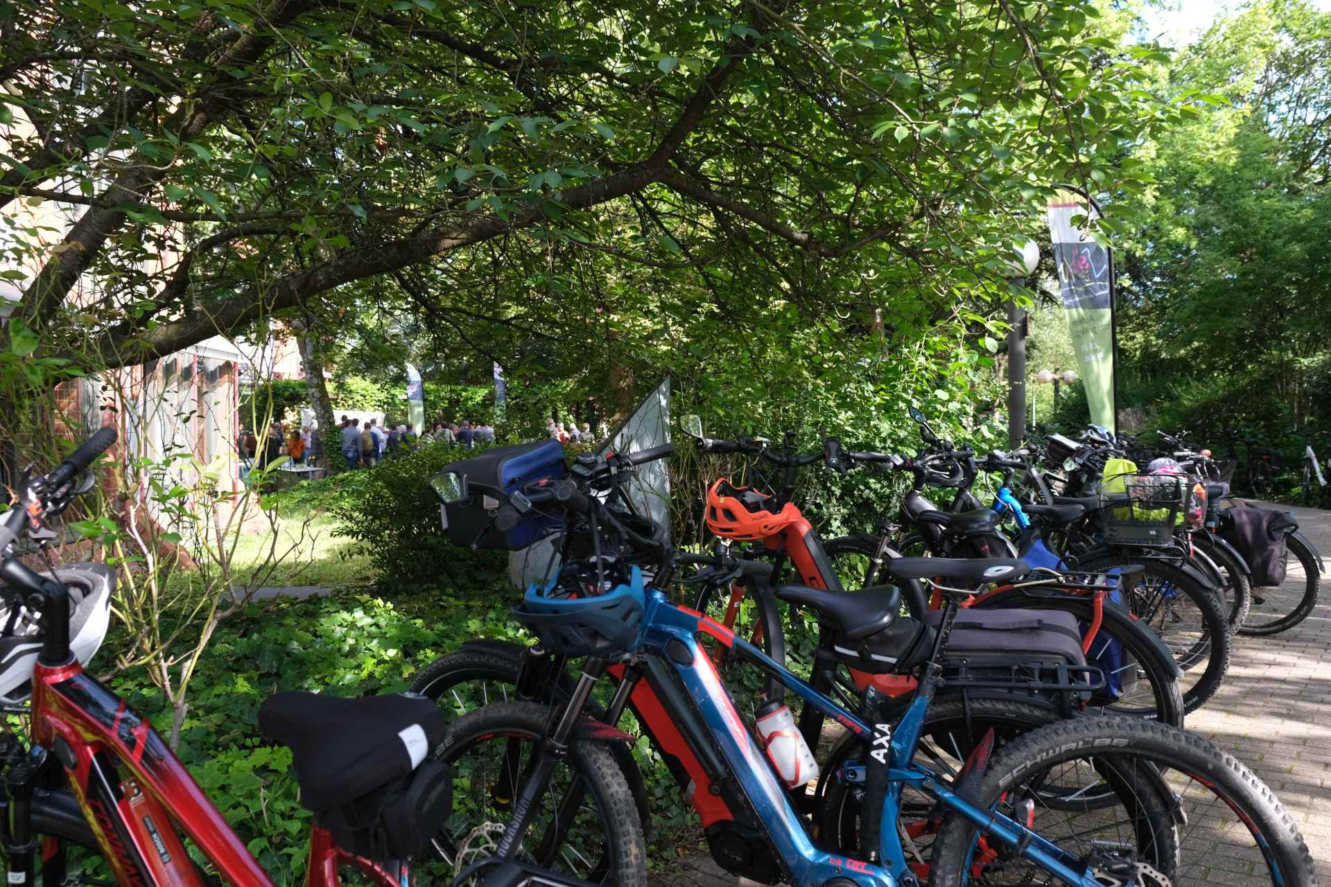 Mehrere Fahrräder, darunter viele E-Bikes, stehen dicht aneinandergereiht an einem Fahrradparkplatz im Grünen. Die Räder sind mit verschiedenen Taschen, Körben und Helmen ausgestattet. Im Hintergrund, unter Bäumen, versammeln sich zahlreiche Menschen bei einer Veranstaltung. Ein Banner mit einem Fahrradmotiv ist neben dem Weg aufgestellt.