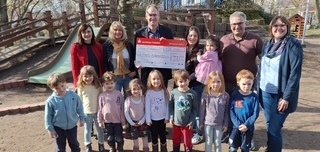 Eine Gruppe aus Erwachsenen und Kindern steht im Außenbereich für ein Foto zusammen und schaut lächelnd in die Kamera. Die Personen in der Mitte halten einen großen symbolischen Scheck. Im Hintergrund sind Bäume und ein Kinderspielplatz mit einer Rutsche zu sehen.