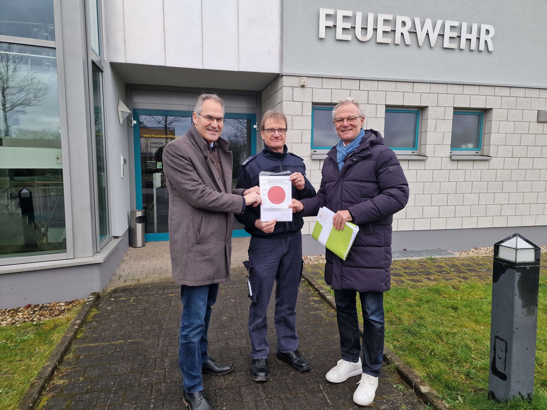 Landrat Dietmar Seefeldt, Wehrleiter Oliver Siebert und VG-Bürgermeister stehen vor dem Feuerwehrhaus in Offenbach, das anhand der Aufschrift "Feuerwehr" deutlich als solches zu erkennen ist. Gemeinsam halten das Baustellenschild mit dem roten Punkt, das die Baufreigabe kennzeichnet.