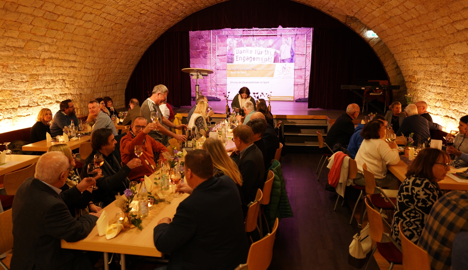 In einem stimmungsvoll beleuchteten Gewölbekeller sitzen zahlreiche Gäste an festlich gedeckten Tischen. Sie stoßen mit Weingläsern an und führen angeregte Gespräche. Im Hintergrund ist eine Bühne mit einer Leinwand zu sehen, auf der die Worte „Danke für Ihr Engagement!“ und „Ehrung der Ehrenamtlichen im Sport“ stehen.