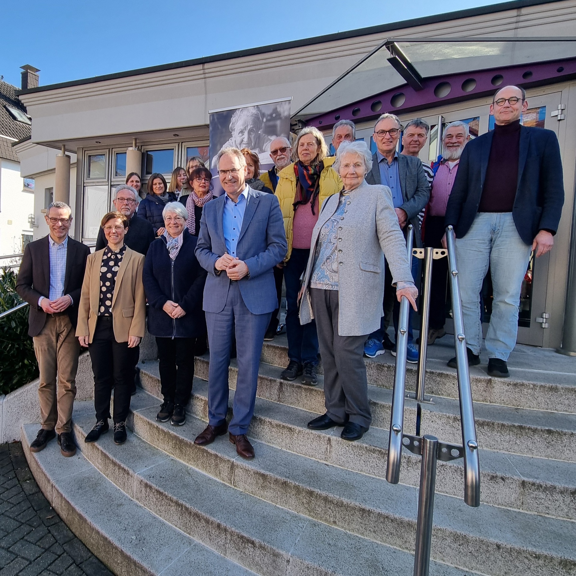 Eine Gruppe von Menschen steht auf der Treppe vor einem Gebäude und blickt in die Kamera. Im Hintergrund ist ein Banner mit einem Porträt zu sehen. Das Foto wurde bei der Eröffnung des Bad Bergzaberner Büros von Silberstreif e.V. aufgenommen.