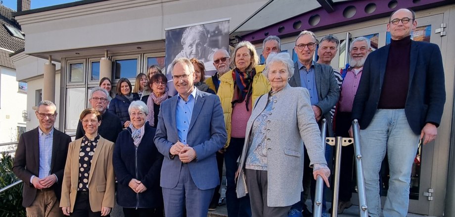 Eine Gruppe von Menschen steht auf der Treppe vor einem Gebäude und blickt in die Kamera. Im Hintergrund ist ein Banner mit einem Porträt zu sehen. Das Foto wurde bei der Eröffnung des Bad Bergzaberner Büros von Silberstreif e.V. aufgenommen.