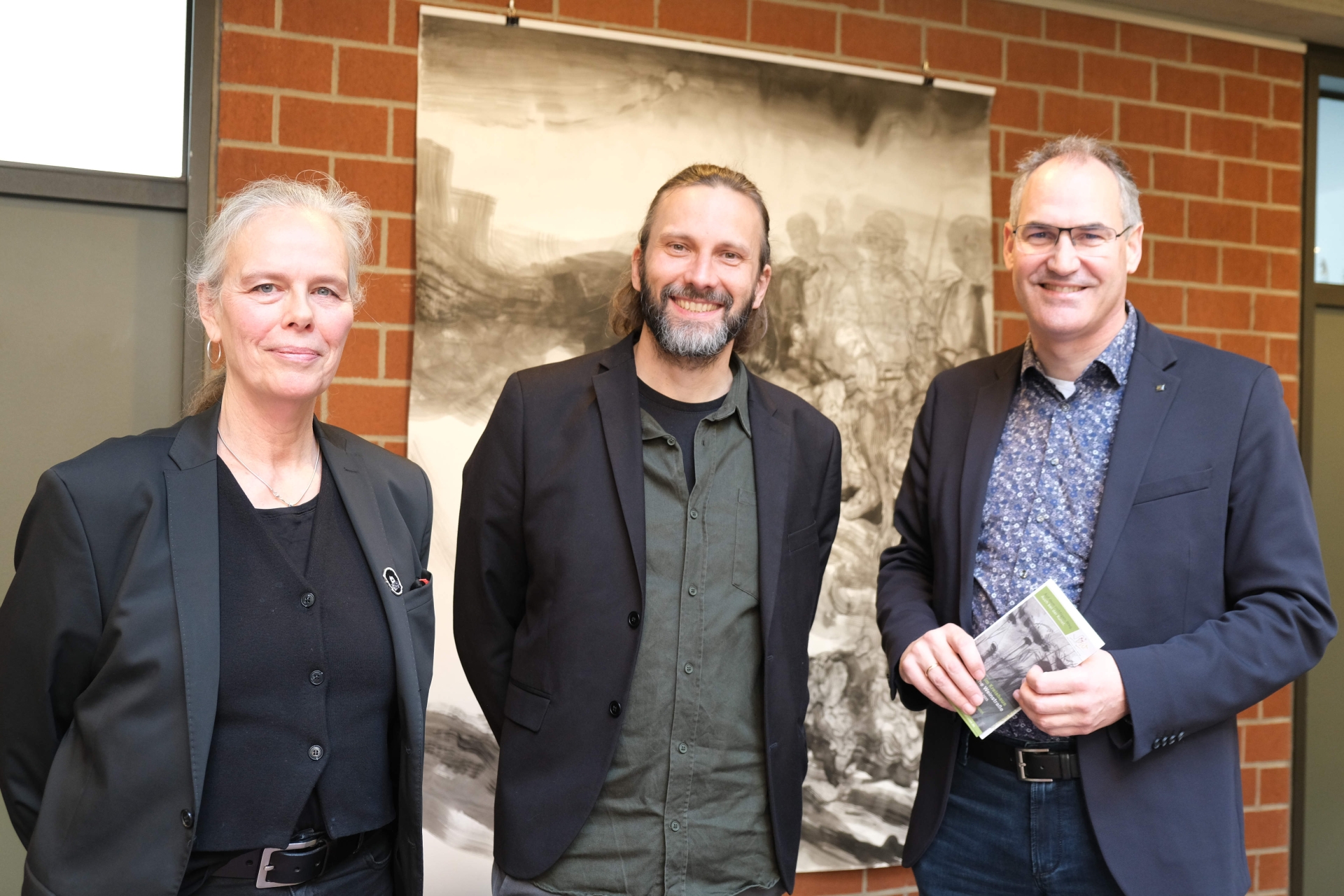 Tina Stolt, Rainer Steve Kaufmann und Dietmar Seefeldt (von links) bei der Ausstellungseröffnung. Foto: KV SÜW Drei Personen stehen nebeneinander vor einer großformatigen Schwarz-Weiß-Zeichnung. Links eine Frau mit zurückgebundenem grauem Haar, schwarzem Blazer und schwarzem Oberteil (Tina Stolt), in der Mitte ein Mann mit schulterlangem Haar und Bart, schwarzer Jacke und olivfarbenem Hemd (Rainer Steve Kaufmann), rechts ein Mann mit Brille, dunklem Blazer und gemustertem Hemd, der eine Broschüre in der Hand hält (Landrat Dietmar Seefeldt). Alle drei blicken in die Kamera und lächeln.