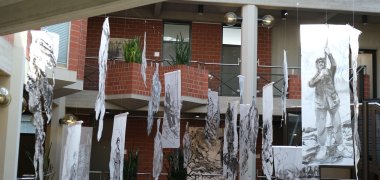 Die preisgekrönte Installation „Tabak“ (2024) steht im Mittelpunkt der Schau. Foto: KV SÜW Kunstinstallation im Kreishaus Südliche Weinstraße, einem offenen, mehrstöckigen Gebäude mit Backsteinwänden und großen Fenstern. Mehrere längliche Bahnen mit schwarz-weißen Zeichnungen von Menschen und Pflanzen hängen von der Decke herab. Die Motive zeigen unter anderem Männer und Frauen bei landwirtschaftlicher Arbeit.