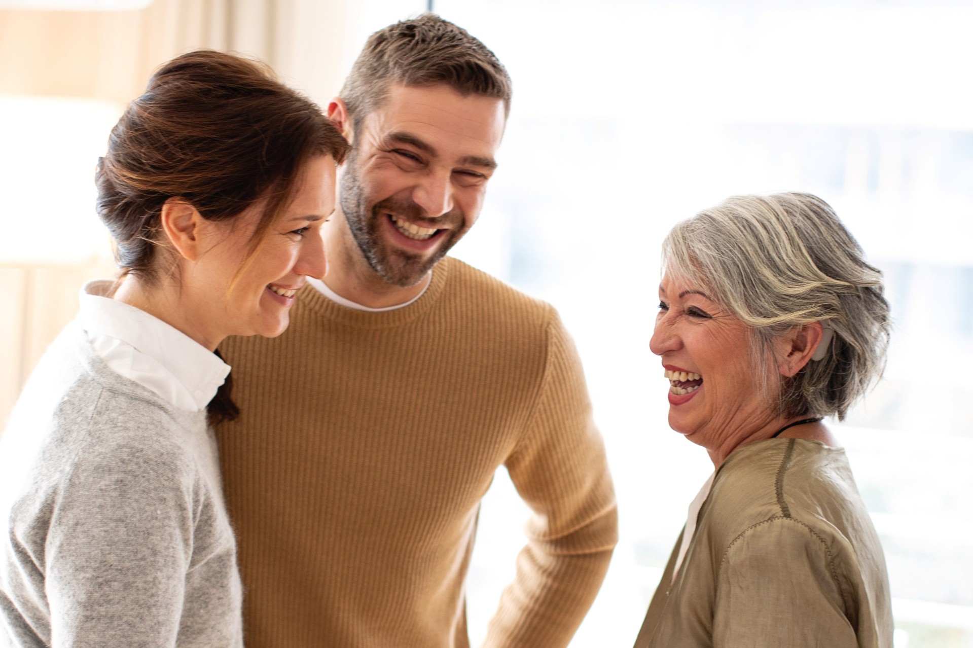 Drei lachende Menschen in einem hellen Raum. Die ältere Frau auf der rechten Seite trägt ein Hörgerät. Das Bild symbolisiert Lebensfreude und zwischenmenschliche Kommunikation.