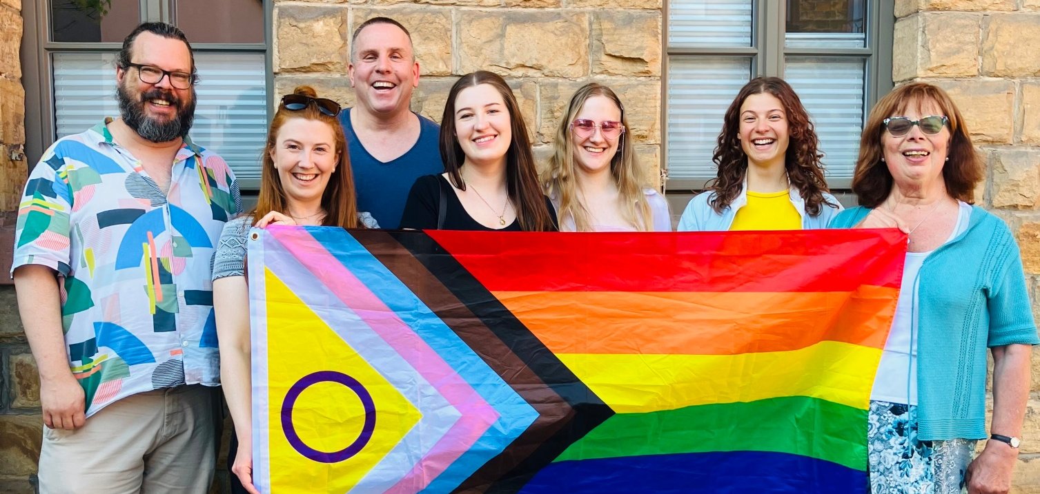Sieben Personen stehen lächelnd vor einer Sandsteinwand und halten gemeinsam die Progress-Pride-Flagge mit Intersektionalitäts-Symbol. Sie zeigen damit ihre Unterstützung für Vielfalt, Akzeptanz und queere Sichtbarkeit in der Südpfalz. Vor ihnen ist mit Kreide eine Sonne auf das Pflaster gemalt.