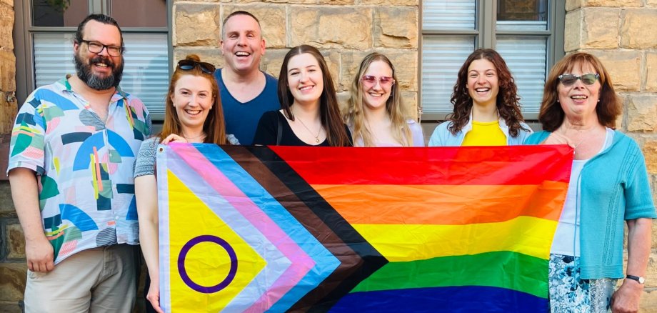 Sieben Personen stehen lächelnd vor einer Sandsteinwand und halten gemeinsam die Progress-Pride-Flagge mit Intersektionalitäts-Symbol. Sie zeigen damit ihre Unterstützung für Vielfalt, Akzeptanz und queere Sichtbarkeit in der Südpfalz. Vor ihnen ist mit Kreide eine Sonne auf das Pflaster gemalt.