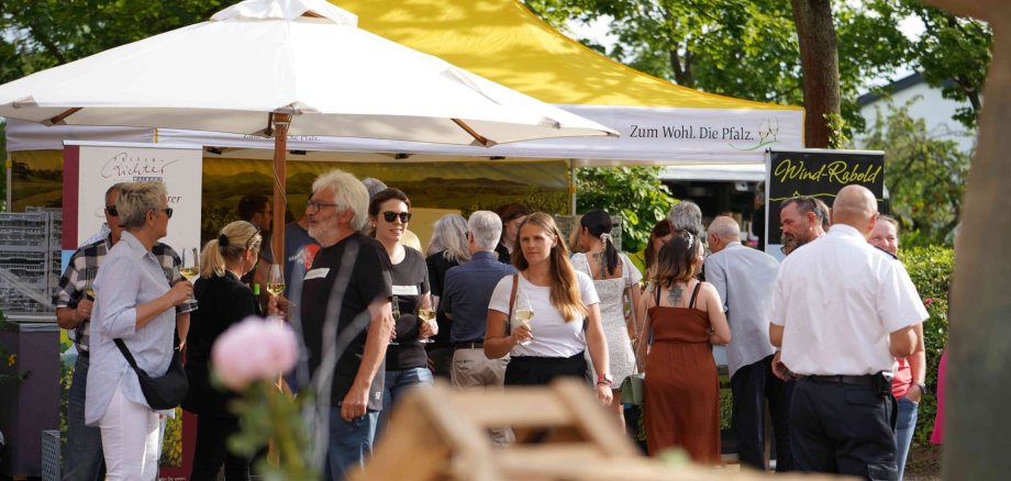 Menschen flanieren mit Stilgläsern in der Hand, im Hintergrund Pagodenzelte, im Vordergrund Weinkisten.
