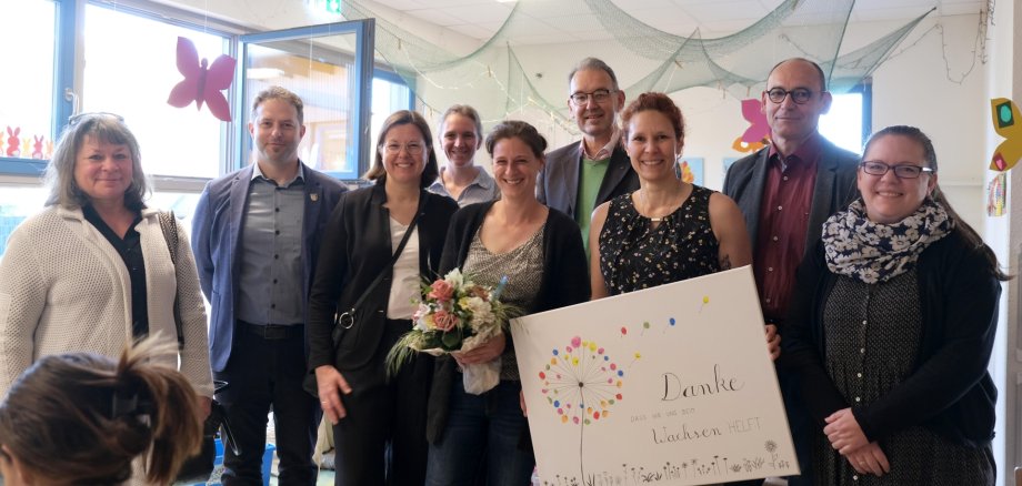 Gruppenbild mit mehreren Frauen und Männern in einem Betreuungsraum der Kta in Rohrbach. Eine Frau hält einen Strauß Blumen, eine weitere ein Plakat mit der Aufschrift "Danke, dass ihr uns beim Wachsen helft". Das Plakat zeigt eine stilisierte Pusteblume in bunten Farben.