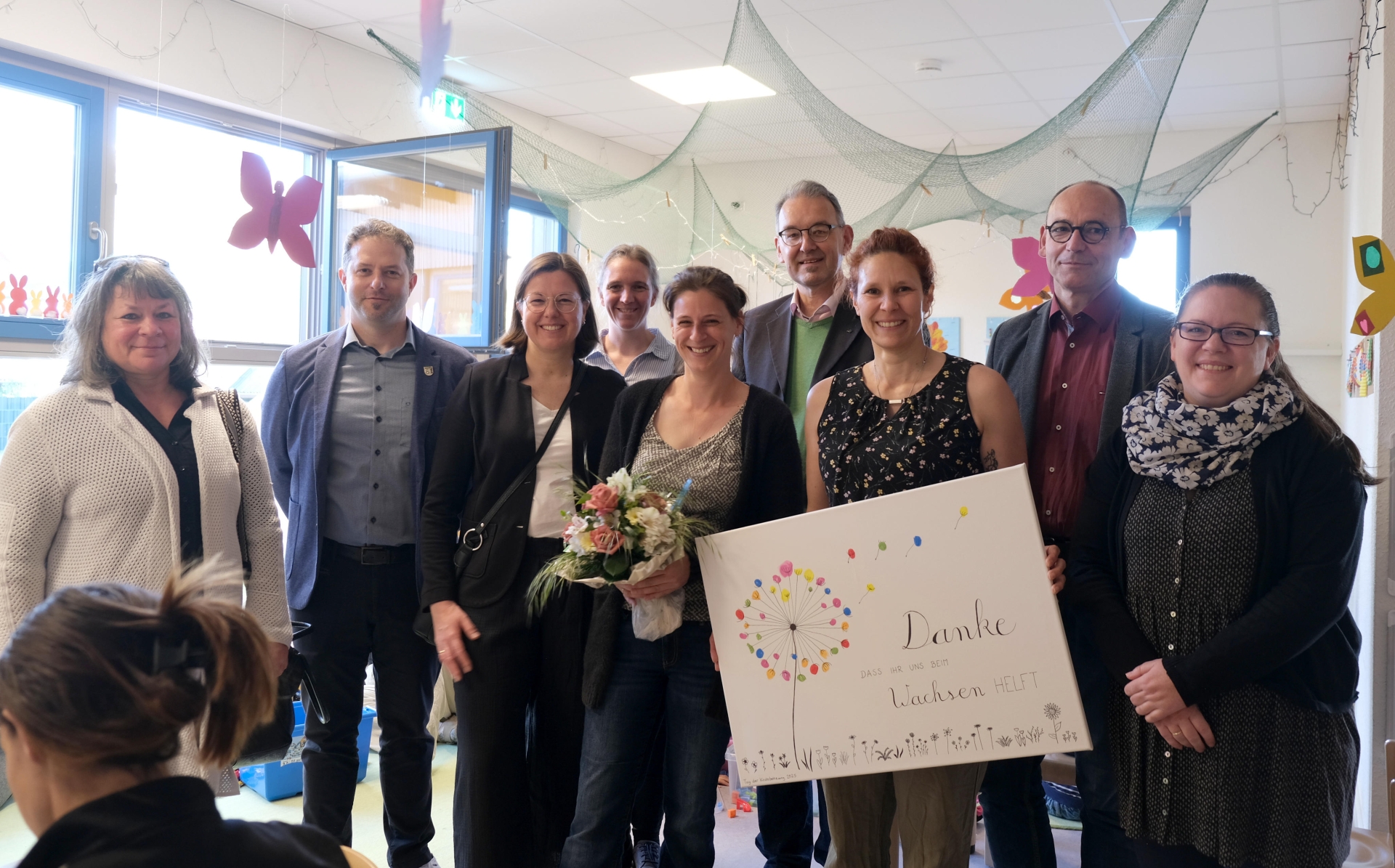 Gruppenbild mit mehreren Frauen und Männern in einem Betreuungsraum der Kta in Rohrbach. Eine Frau hält einen Strauß Blumen, eine weitere ein Plakat mit der Aufschrift "Danke, dass ihr uns beim Wachsen helft". Das Plakat zeigt eine stilisierte Pusteblume in bunten Farben.