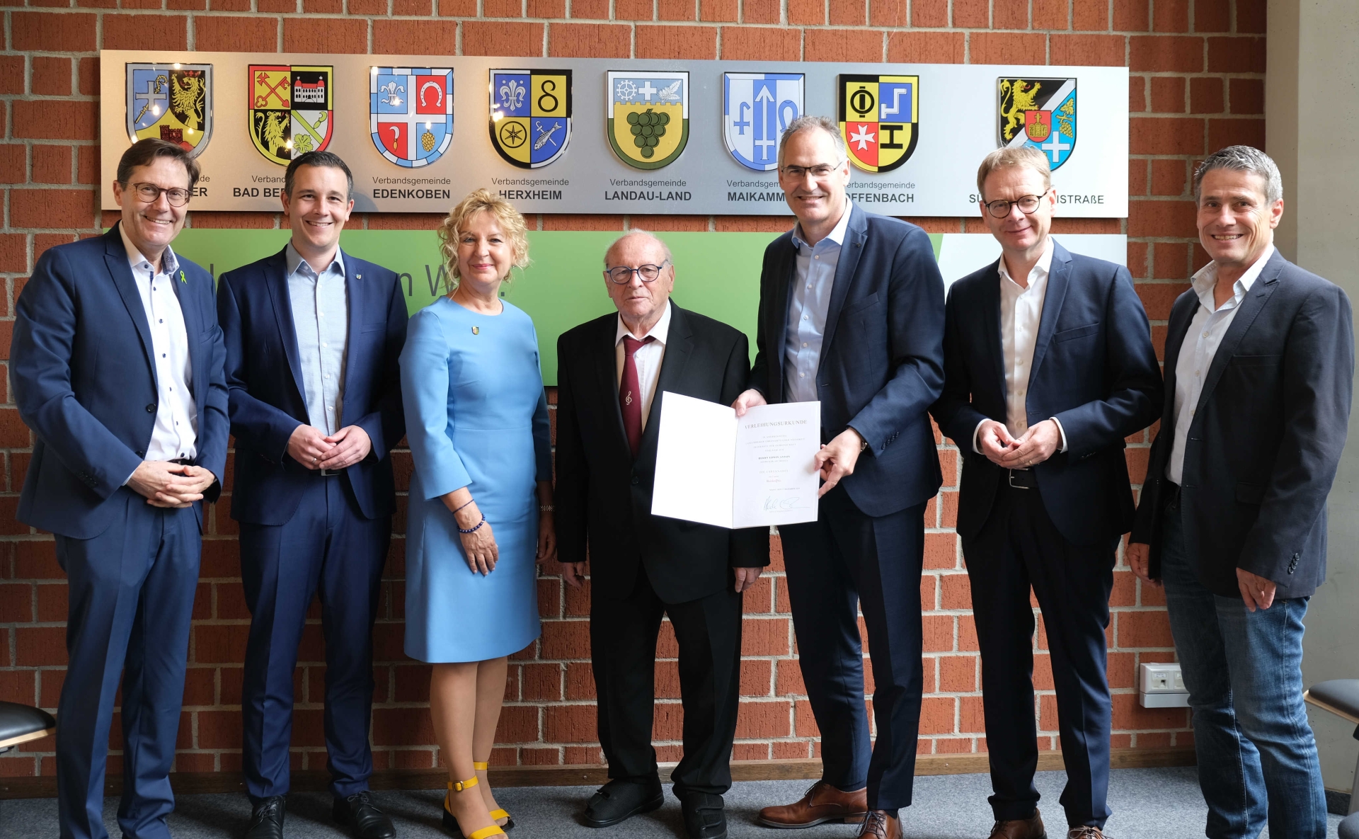 Gruppenfoto: Edwin Anton mit Gratulanten bei der Ehrungsfeier im Kreishaus. Insgesamt sieben Personen stehen vor einer Backsteinwand, an der die Wappen der zum Landkreis SÜW gehörenden Verbandsgemeinden sowie des Landkreises selbst zu sehen sind.
