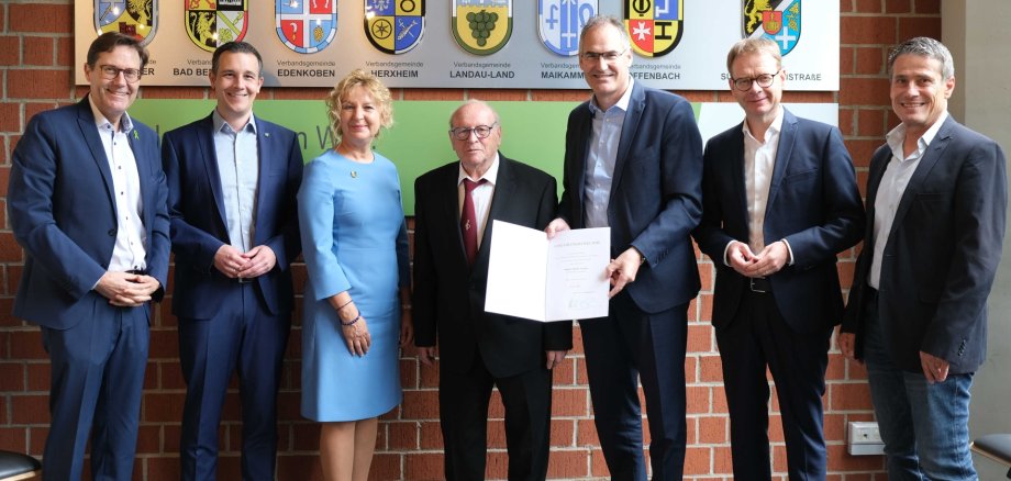Edwin Anton (Mitte) im Kreis der Gratulanten bei der Ehrungsfeier im Kreishaus. Foto KV SÜW Gruppenfoto: Edwin Anton mit Gratulanten bei der Ehrungsfeier im Kreishaus. Insgesamt sieben Personen stehen vor einer Backsteinwand, an der die Wappen der zum Landkreis SÜW gehörenden Verbandsgemeinden sowie des Landkreises selbst zu sehen sind.