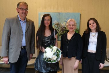 Von links nach rechts: Georg Kern, Judith Boy, Christine Schön und Musikerin Lilly Sophie Reiß, die die Ausstellungseröffnung mit Klavier, Ukulele und Gesang begleitete. Foto: KV SÜW Vier Personen stehen nebeneinander und lächeln in die Kamera. Von links nach rechts: Georg Kern, Judith Boy, Christine Schön und Musikerin Lilly Sophie Reiß. Im Hintergrund sind zwei farbige Gemälde zu sehen.