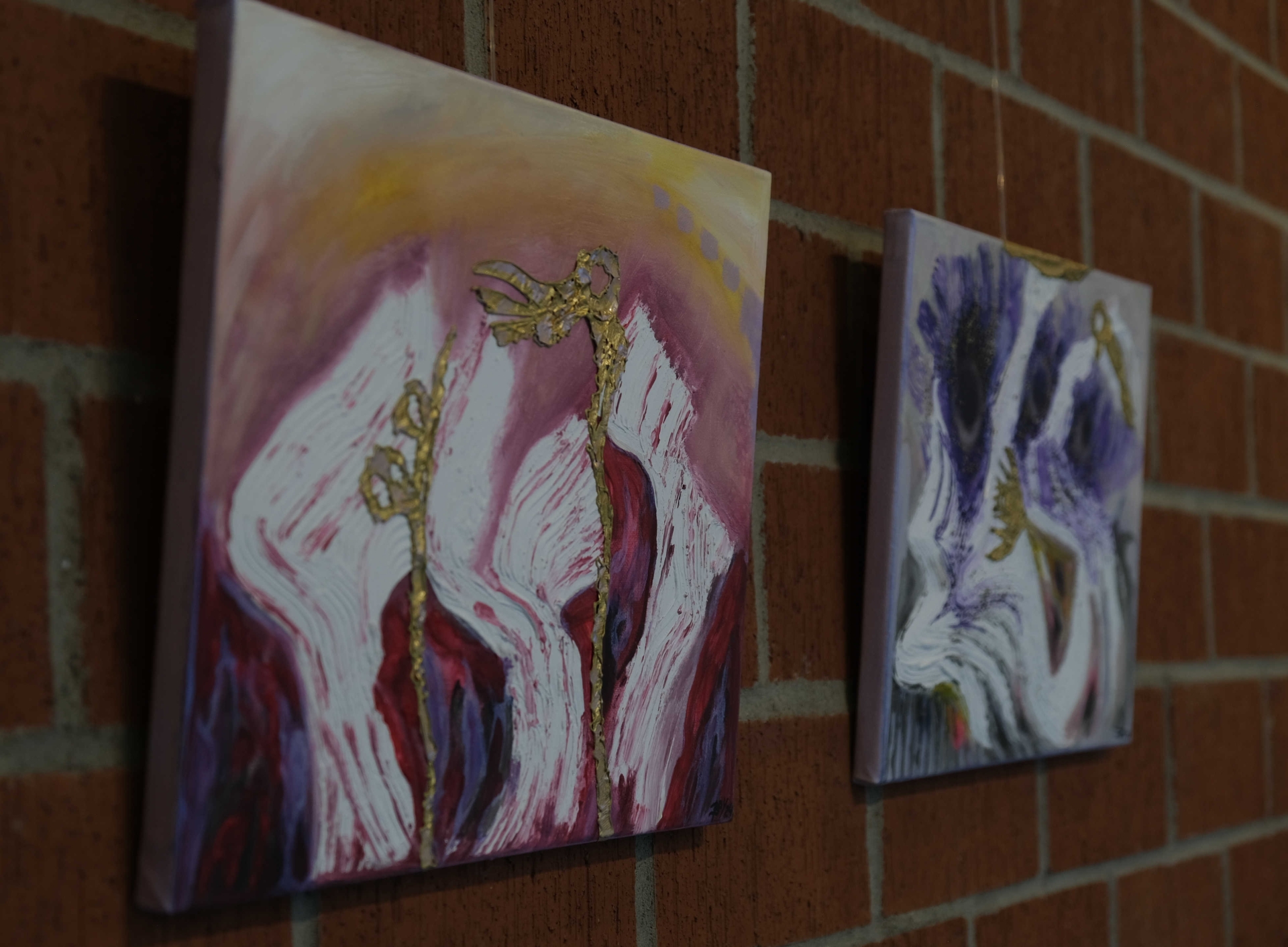Zwei farbintensive, abstrakte Gemälde mit kräftigen Linien und goldenen Strukturen hängen an einer Ziegelwand. Die Farben reichen von Rosa und Rot bis zu Violett und Weiß.