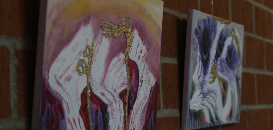 Zwei der Werke von Judith Boy, die derzeit im Kreishaus zu bewundern sind. Foto: KV SÜW Zwei farbintensive, abstrakte Gemälde mit kräftigen Linien und goldenen Strukturen hängen an einer Ziegelwand. Die Farben reichen von Rosa und Rot bis zu Violett und Weiß.