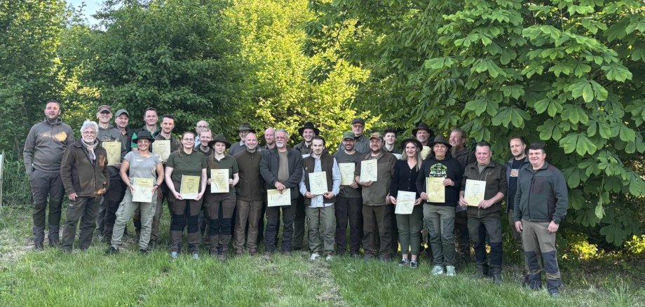 Eine Gruppe von rund 30 Personen steht auf einer Wiese vor dichtem, grünem Laub. Viele von ihnen halten Urkunden in den Händen. Alle tragen outdoor- oder jagdtypische Kleidung und lächeln in die Kamera.