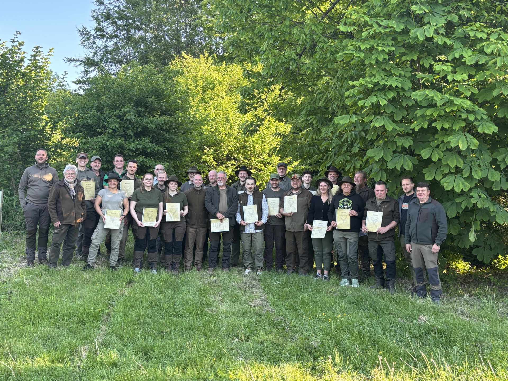 Eine Gruppe von rund 30 Personen steht auf einer Wiese vor dichtem, grünem Laub. Viele von ihnen halten Urkunden in den Händen. Alle tragen outdoor- oder jagdtypische Kleidung und lächeln in die Kamera.