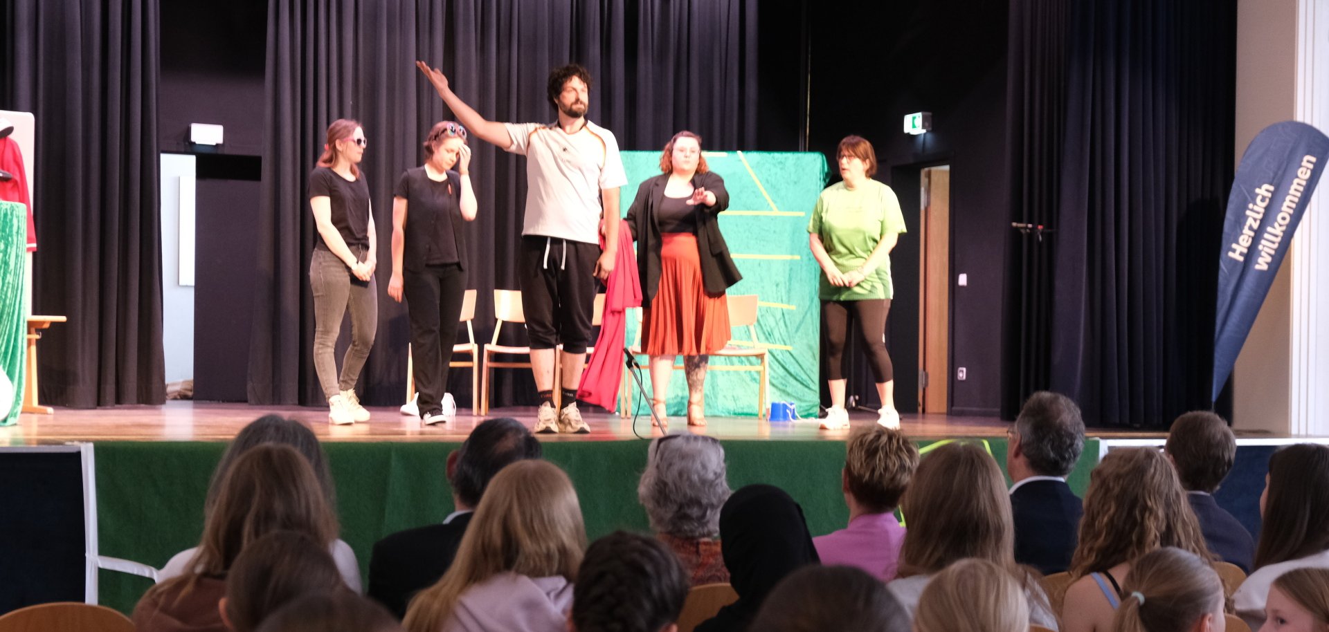 Eine Theatergruppe mit fünf Darstellerinnen und Darstellern steht auf einer Bühne und spricht in Richtung Publikum. Im Zuschauerraum sitzen Kinder und Erwachsene, einige tragen Halstücher mit Malteser-Emblem. Rechts am Bühnenrand steht ein Aufsteller mit der Aufschrift ‚Herzlich willkommen‘.