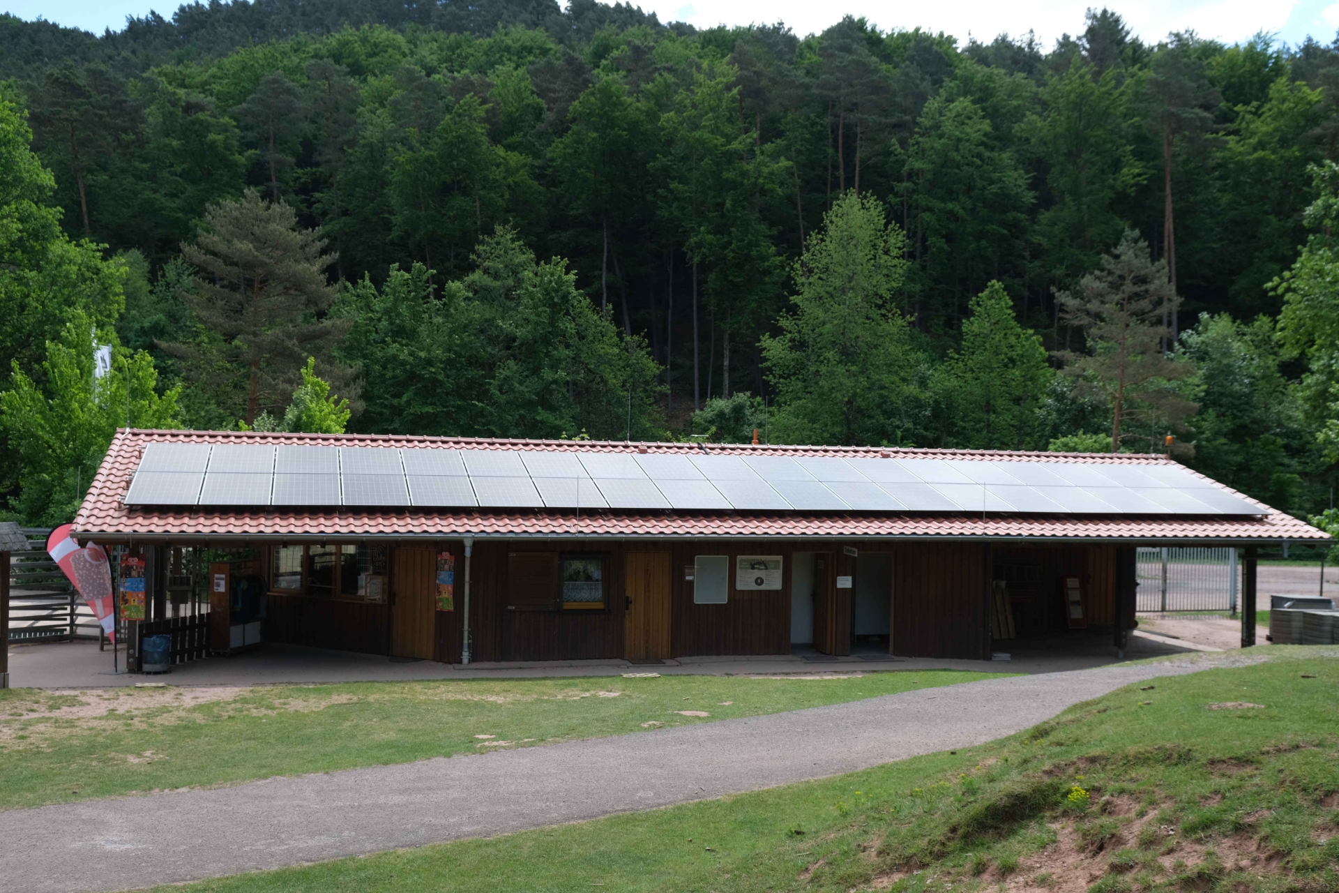 Ein langgezogenes Holzgebäude mit rotem Ziegeldach und großen Photovoltaik-Modulen auf dem Dach. Umgeben ist das Gebäude von Wald und Wiese im Wild- und Wanderpark in Silz.