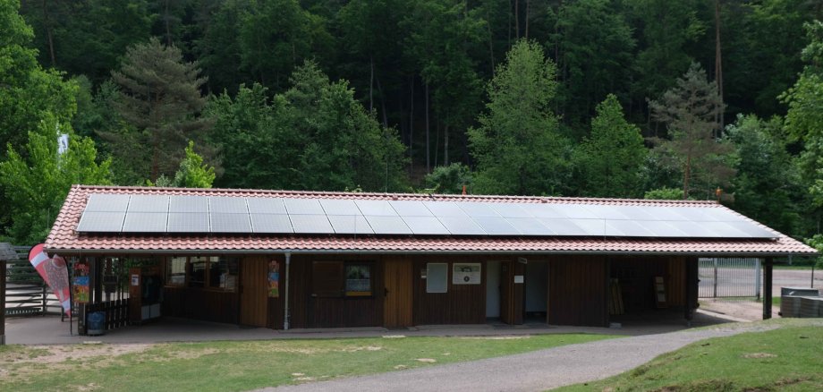 Ein langgezogenes Holzgebäude mit rotem Ziegeldach und großen Photovoltaik-Modulen auf dem Dach. Umgeben ist das Gebäude von Wald und Wiese im Wild- und Wanderpark in Silz.