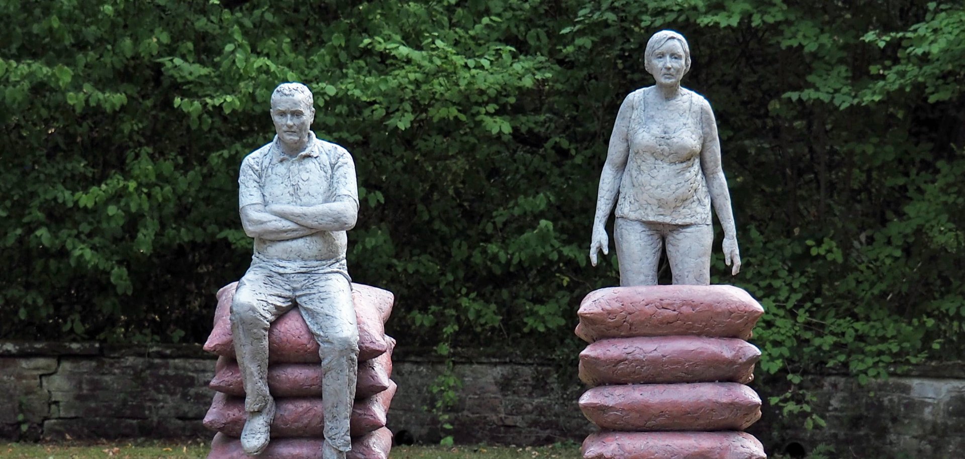 Zwei Betonskulpturen zeigen eine Frau und einen Mann, die auf hohen Stapeln aus pinkfarbenen, kissenartigen Blöcken stehen. Der Mann sitzt mit verschränkten Armen, die Frau steht aufrecht.