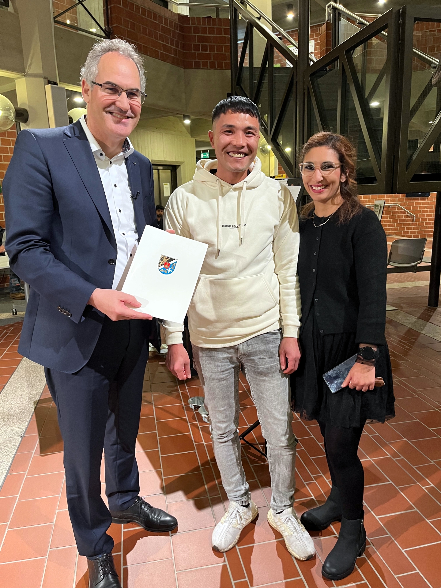 Landrat Dietmar Seefeldt überreicht im Foyer des Kreishauses der Südlichen Weinstraße Javad Alizadeh aus Afghanistan die Einbürgerungskunde, rechts die Integrationsbeauftragte des Landkreises Südliche Weinstraße Sihame Hlubek.