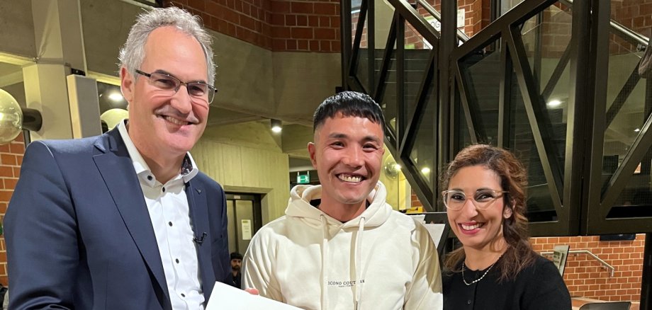 Landrat Dietmar Seefeldt überreicht im Foyer des Kreishauses der Südlichen Weinstraße Javad Alizadeh aus Afghanistan die Einbürgerungskunde, rechts die Integrationsbeauftragte des Landkreises Südliche Weinstraße Sihame Hlubek.