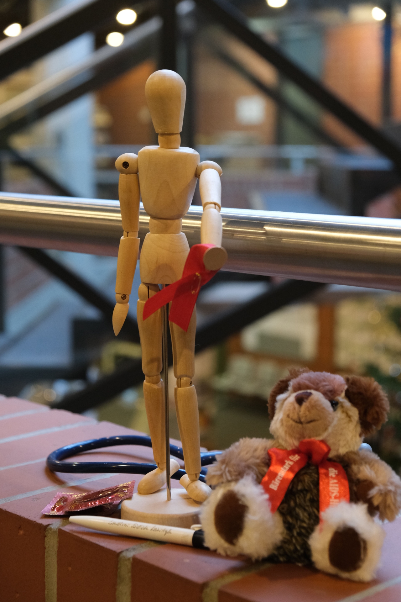 Ein Männchen aus Holz hält eine rote Schleife in der Hand, davor ein Kugelschreiber der Kreisverwaltung SÜW, ein Kondom und ein Plüschbär der AIDS-Hilfe.